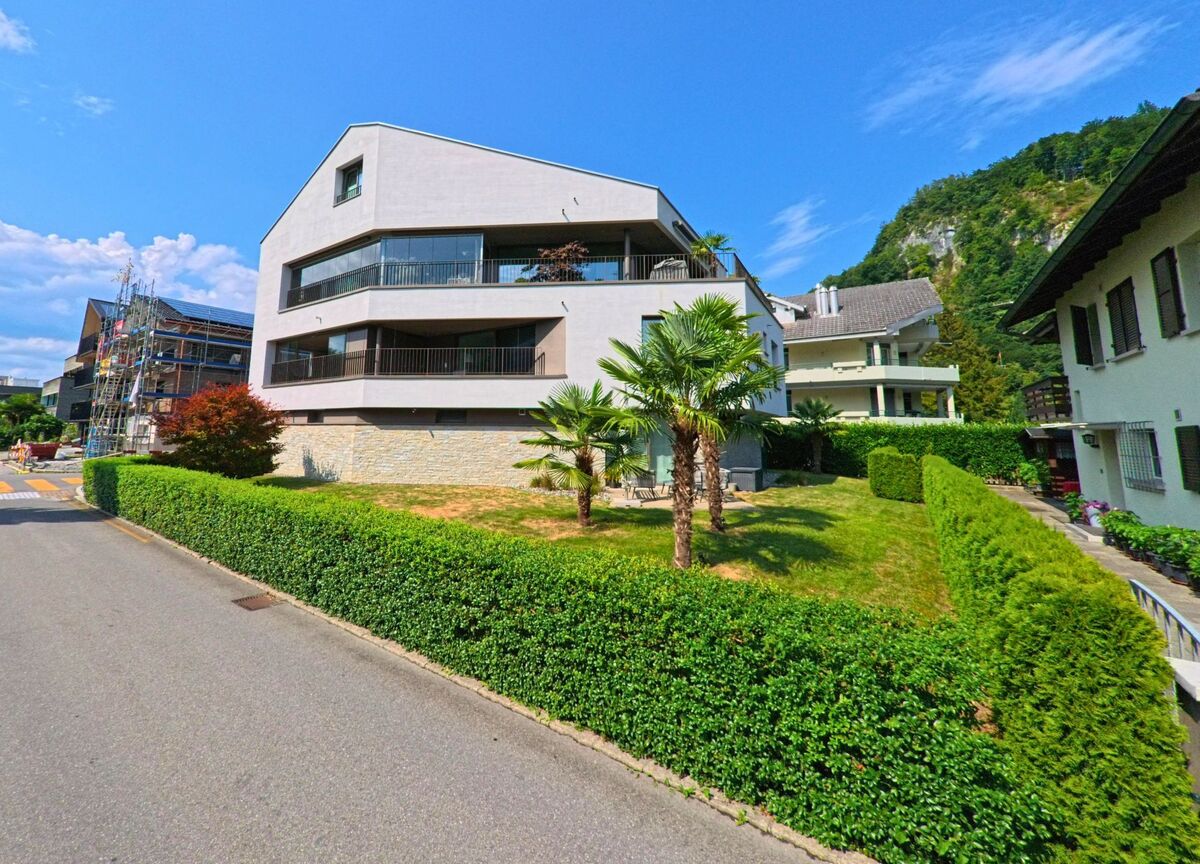 In einem Wohnquartier von Stansstad (6362, Kanton Nidwalden) steht ein modernes Mehrfamilienhaus mit markanter Fassade und Palmen im gepflegten Vorgarten vor einer bewaldeten Hangkulisse.