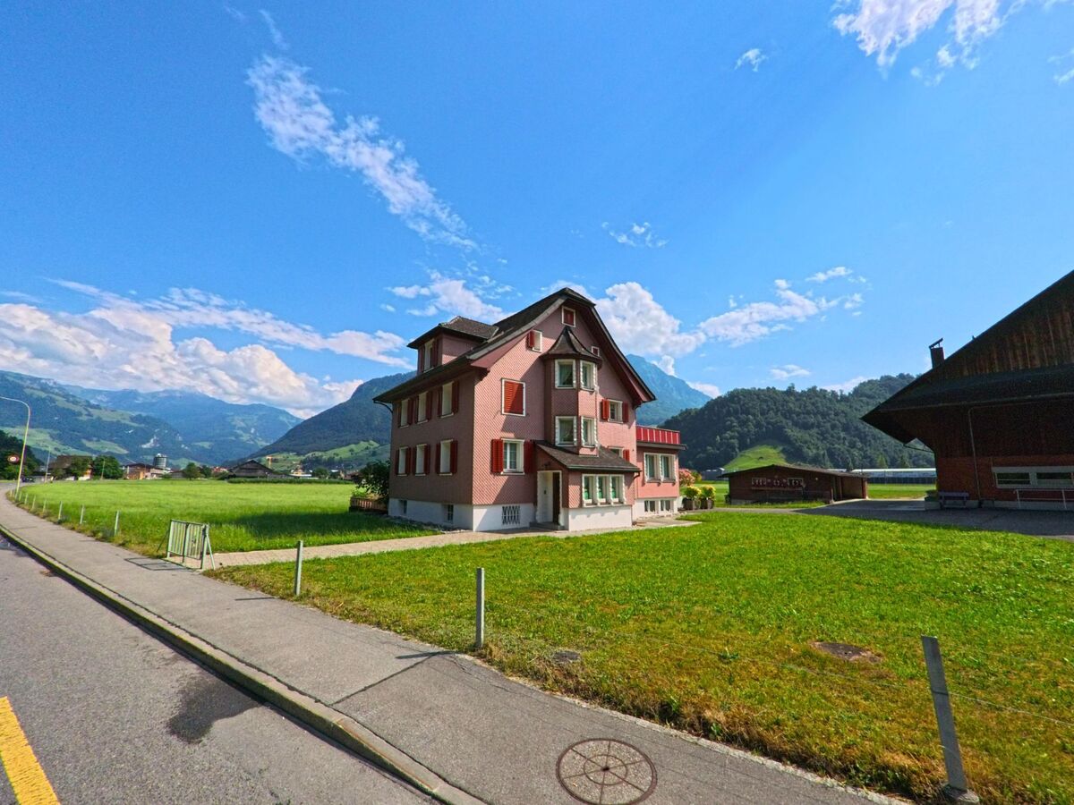 Am Ortsrand von Stansstad (6362, Kanton Nidwalden) steht ein rotes Wohnhaus mit Fensterlaeden auf einer Wiese, umgeben von Bergen und traditioneller Bebauung.
