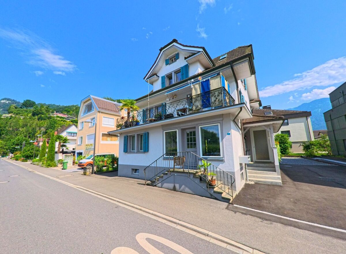 In einer ruhigen Wohnstrasse von Stansstad (6362, Kanton Nidwalden) steht ein weiss gestrichenes Einfamilienhaus mit blauen Fensterläden und verziertem Balkon vor grüner Hügellandschaft.