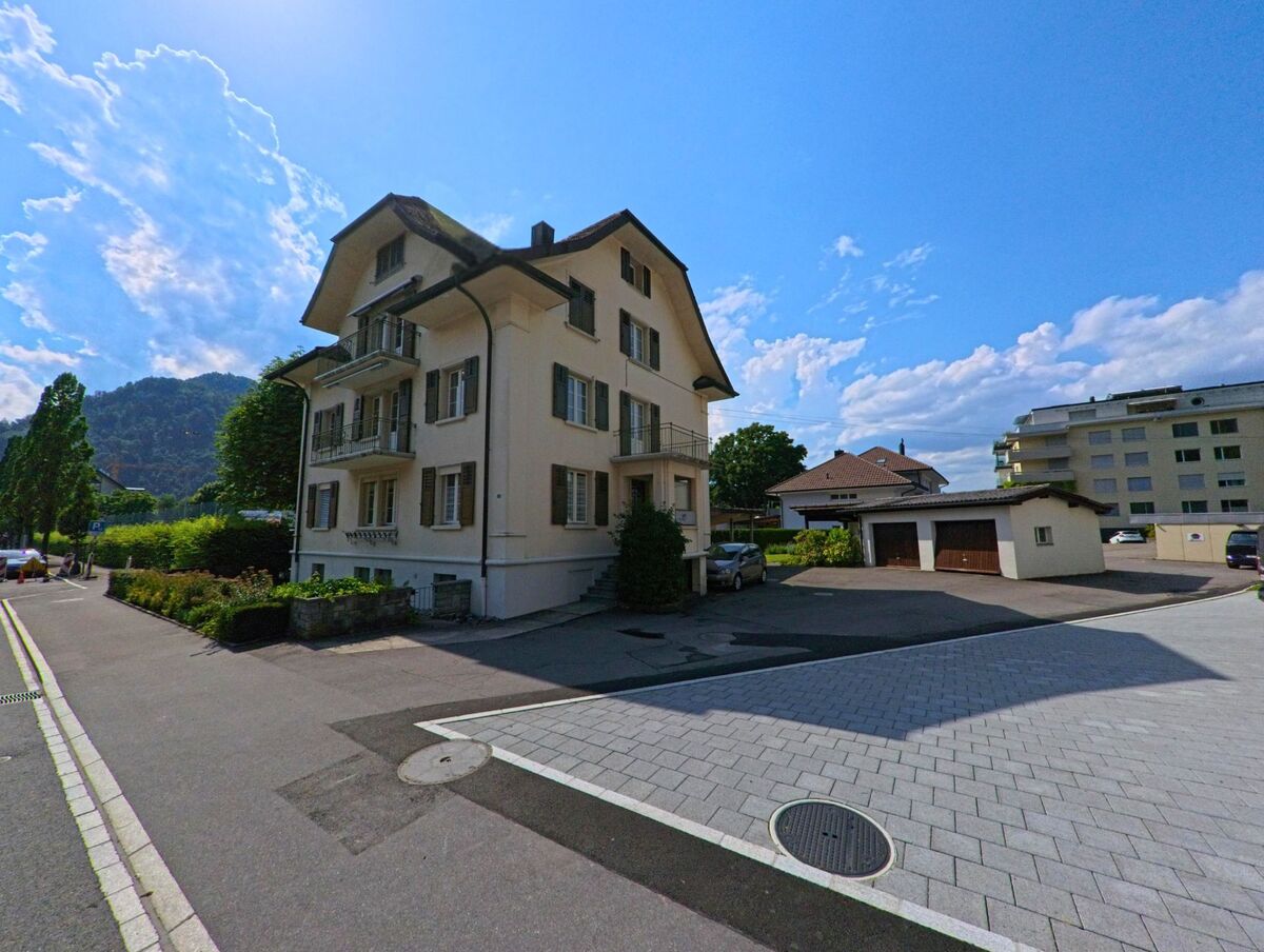 In einem Wohngebiet von Stansstad (6362, Kanton Nidwalden) steht ein mehrstöckiges, helles Wohnhaus mit gruenen Fensterlaeden und Balkonen, umgeben von Garagen und modernerer Bebauung.