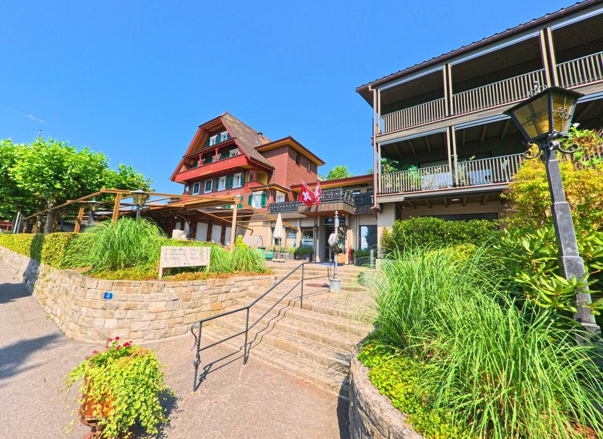 Eine Anlage mit traditionell und modern gestalteten Gebaeuden in Stansstad (6363, Kanton Nidwalden) wird von gepflegten Terrassen, Bepflanzung und einer Schweizer Fahne am Eingangsausschnitt ergänzt.