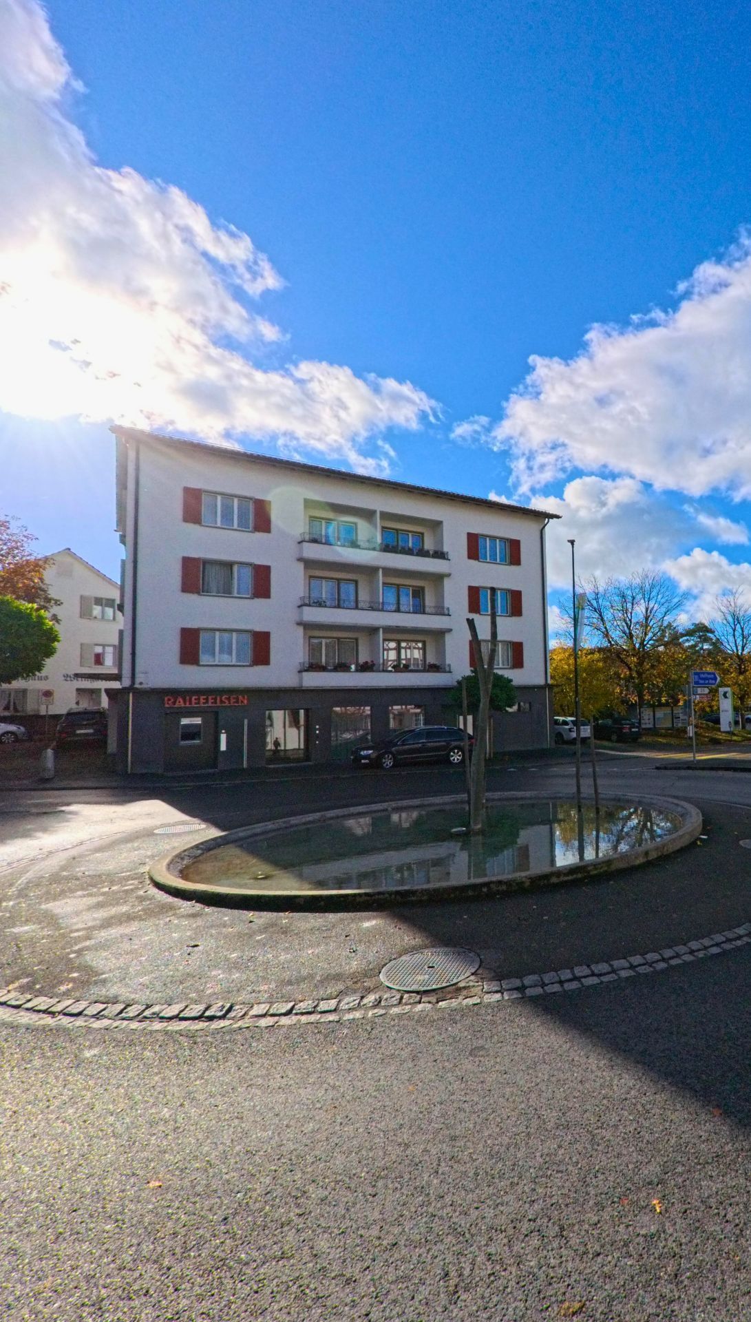 An einem kleinen Kreisplatz mit Wasserbecken steht in Steckborn (8266, Kanton Thurgau) ein Wohn- und Geschaeftshaus unter blauem Himmel mit vorbeiziehenden Wolken.
