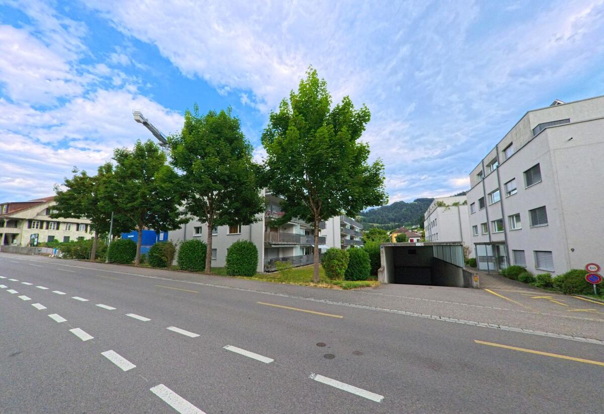 In Steffisburg (3612, Kanton Bern) saeumen moderne Wohnblocks mit Tiefgaragenzufahrt eine mehrspurige Strasse, zwischen den Gebaeuden stehen gleichmaessig gepflanzte Baeume.
