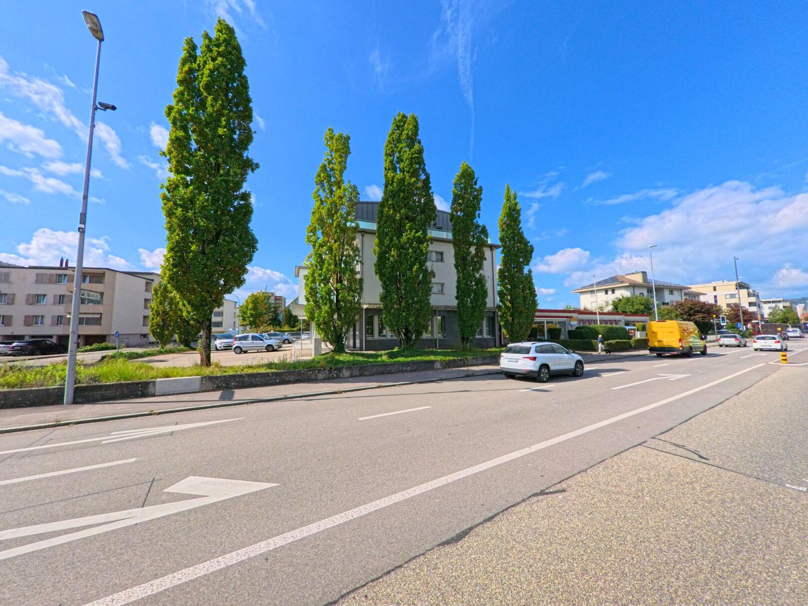 Entlang einer belebten Strasse in Stein (4332, Kanton Aargau) stehen hohe, schlanke Baume vor einem modernen Gebaude, waehrend mehrere Fahrzeuge vorbeifahren.