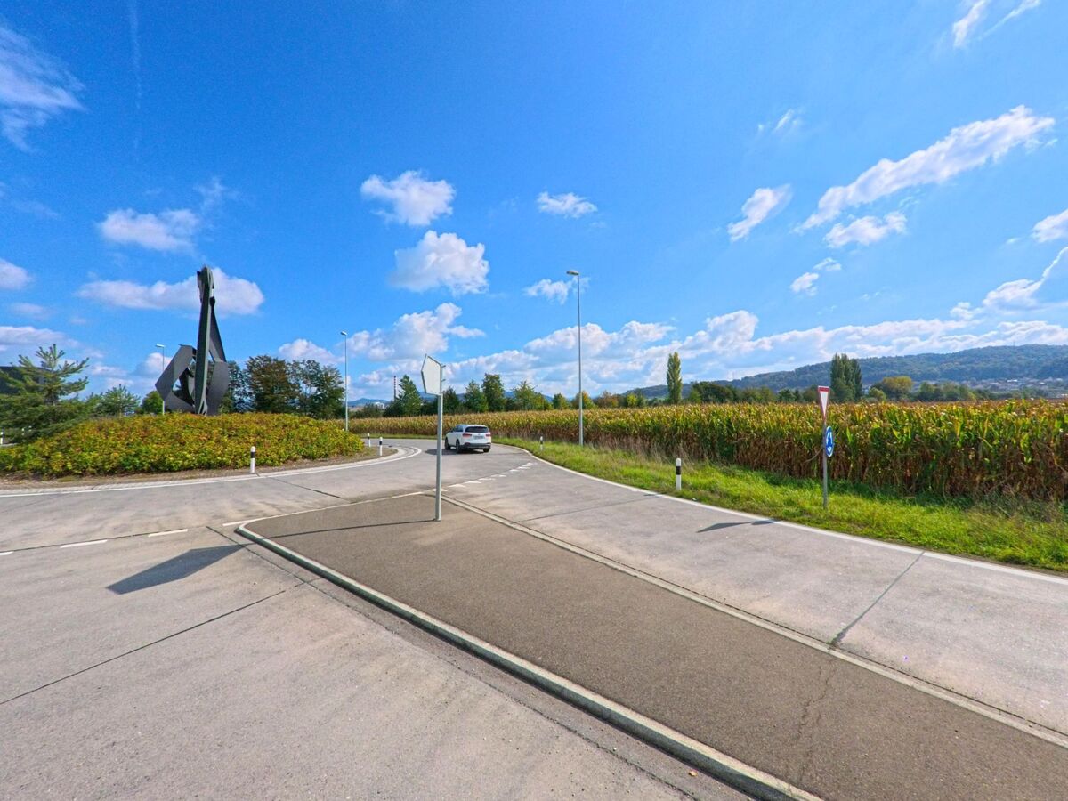 Am Rand von Stein (4332, Kanton Aargau) fuehrt eine Strasse an einem Kreisverkehr mit moderner Skulptur vorbei, umgeben von Feldern und einer weiten Huegellandschaft.