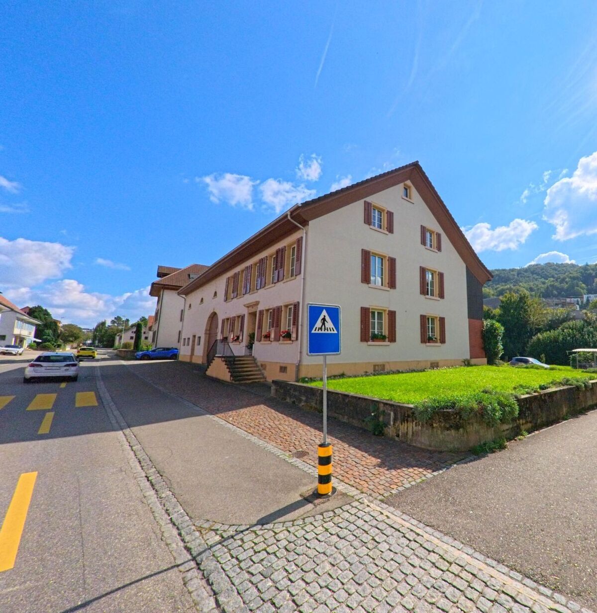 Ein traditionelles Gebaeude mit braunen Fensterlaeden praegt in Stein (4332, Kanton Aargau) eine leicht ansteigende Strasse, die von gepflasterten Bereichen und Gruenflaechen begleitet wird.