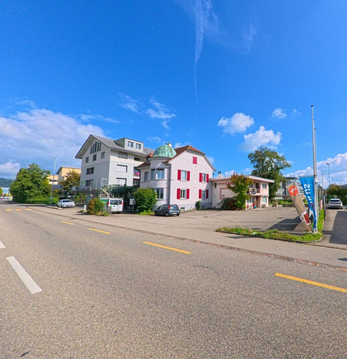 In Stein (4332, Kanton Aargau) stehen entlang der Hauptstrasse ein helles Wohnhaus mit markanten roten Fensterlaeden und ein kleines Cafe mit Parkplatz davor.