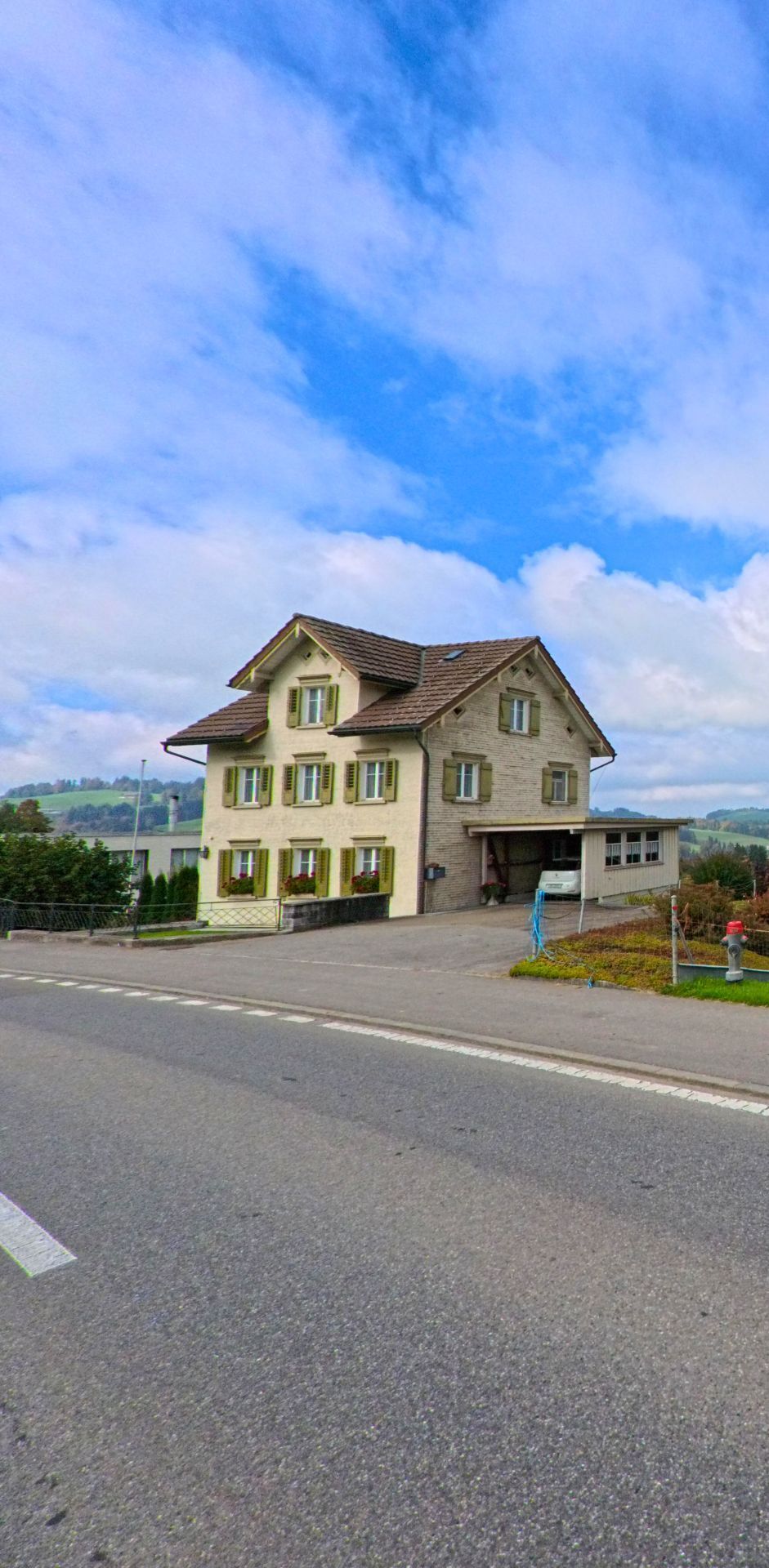 In Stein (9063, Kanton Appenzell Ausserrhoden) steht ein helles Wohnhaus mit grünen Fensterlaeden leicht erhoeht an einer Landstrasse mit weitem Blick ins Hügelland.