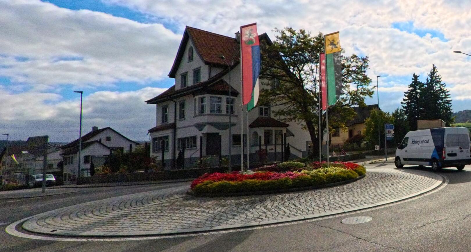 Vor einem Wohnhaus in Stein am Rhein (8260, Kanton Schaffhausen) zeigt ein bepflanzter Kreisel mit zwei Fahnen eine gepflegte Verkehrsinsel unter leicht bewolktem Himmel.