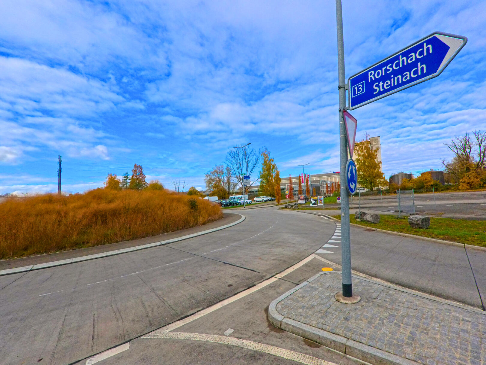 An einer Verkehrsinsel in Steinach (9323, Kanton St. Gallen) zeigt ein blaues Wegweisserschild die Richtung nach Rorschach unter wolkigem Himmel.