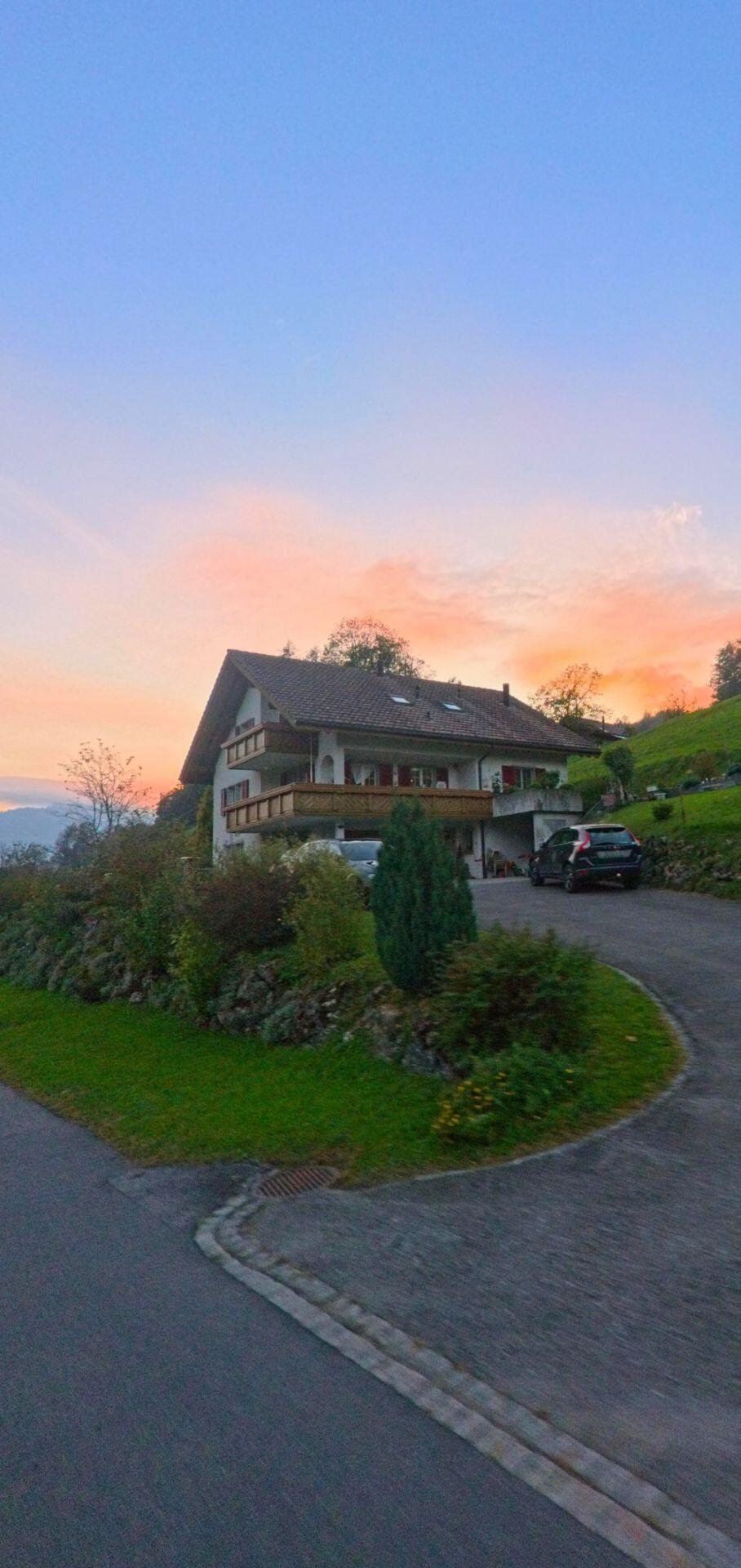 Vor einem farbigen Abendhimmel steht in Steinen (6422, Kanton Schwyz) ein Haus mit grossem Balkon an einem leicht ansteigenden Hang, umgeben von Gartenpflanzen und einer Zufahrt.
