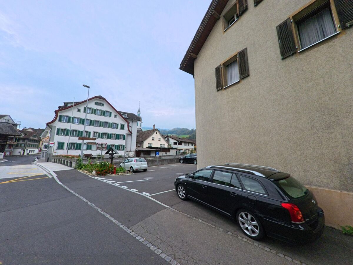 Im Zentrum von Steinen (6422, Kanton Schwyz) stehen traditionelle Wohnhaeuser mit gruenen Fensterlaeden, waehrend im Vordergrund ein Parkplatz und klare Strassenmarkierungen sichtbar sind.
