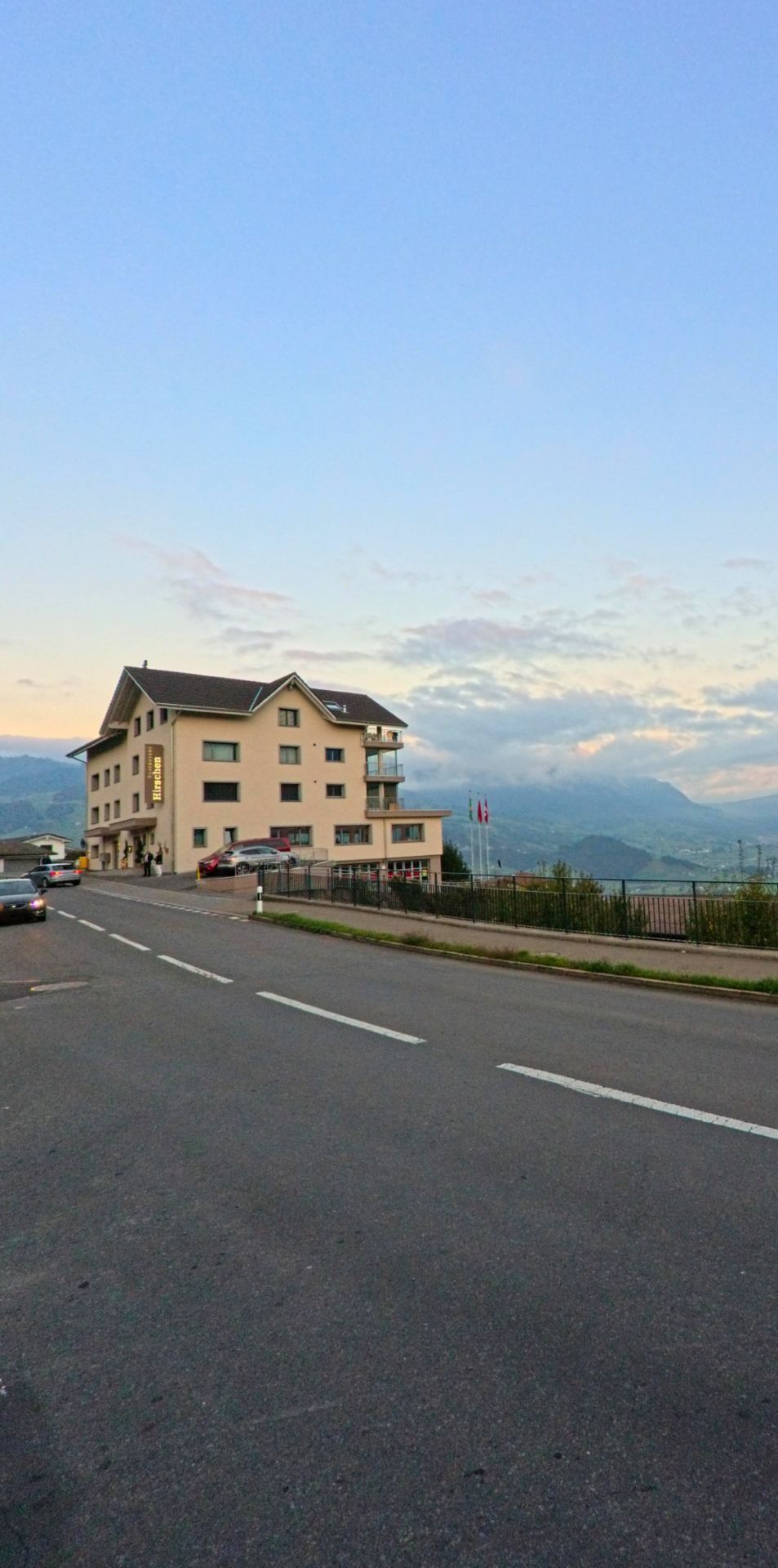 Ein mehrstockiges Gebaeude in Steinerberg (6416, Kanton Schwyz) steht an einer ansteigenden Strasse, waehrend sich dahinter eine weite Berglandschaft unter einem hellen Abendhimmel ausbreitet.