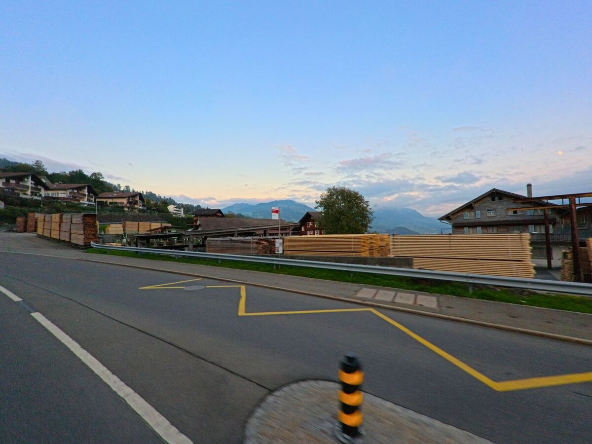 In Steinerberg (6416, Kanton Schwyz) liegen gestapelte Holzbohlen entlang einer Strasse, waehrend dahinter Wohnhaeuser und eine weite Bergkulisse im Abendlicht sichtbar sind.