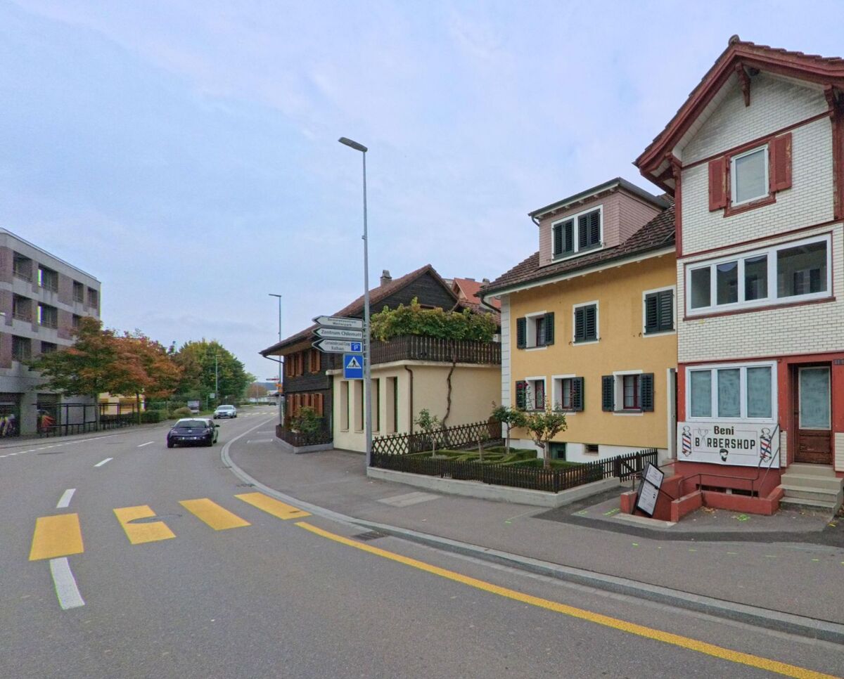 Entlang einer leicht gebogenen Strasse in Steinhausen (6312, Kanton Zug) stehen Wohnhaeuser mit Fensterlaeden und kleinen Gaerten nahe einem Fussgaengeruebergang.