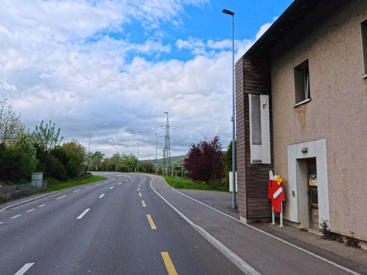 Eine breite Strasse mit Mittelstreifen führt in Steinmaur (8162, Kanton Zürich) an einem schlichten Gebäude vorbei in Richtung eines von Bäumen gesäumten Horizonts.
