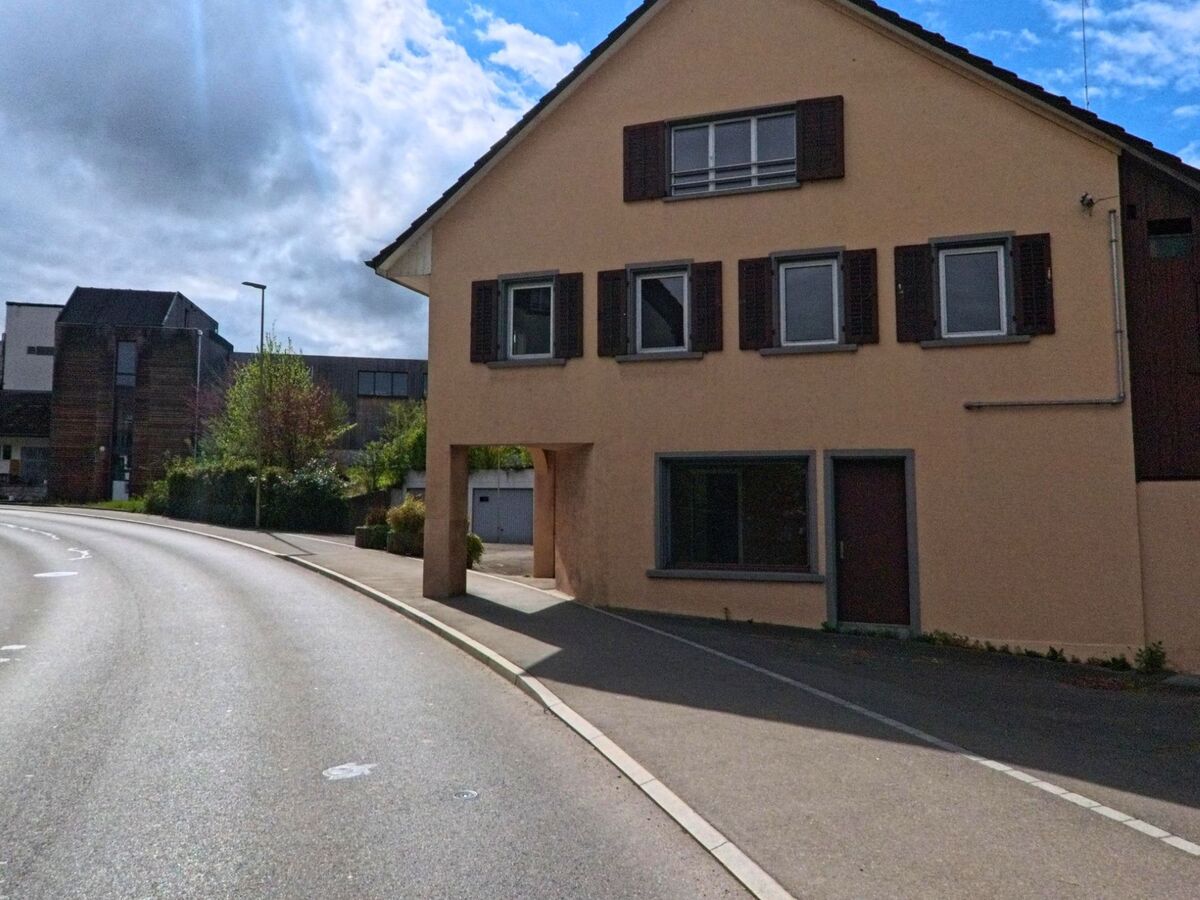 In Steinmaur (8162, Kanton Zürich) führt eine sanft gebogene Strasse an einem Wohnhaus mit Torbogen vorbei, das teilweise im Schatten liegt.
