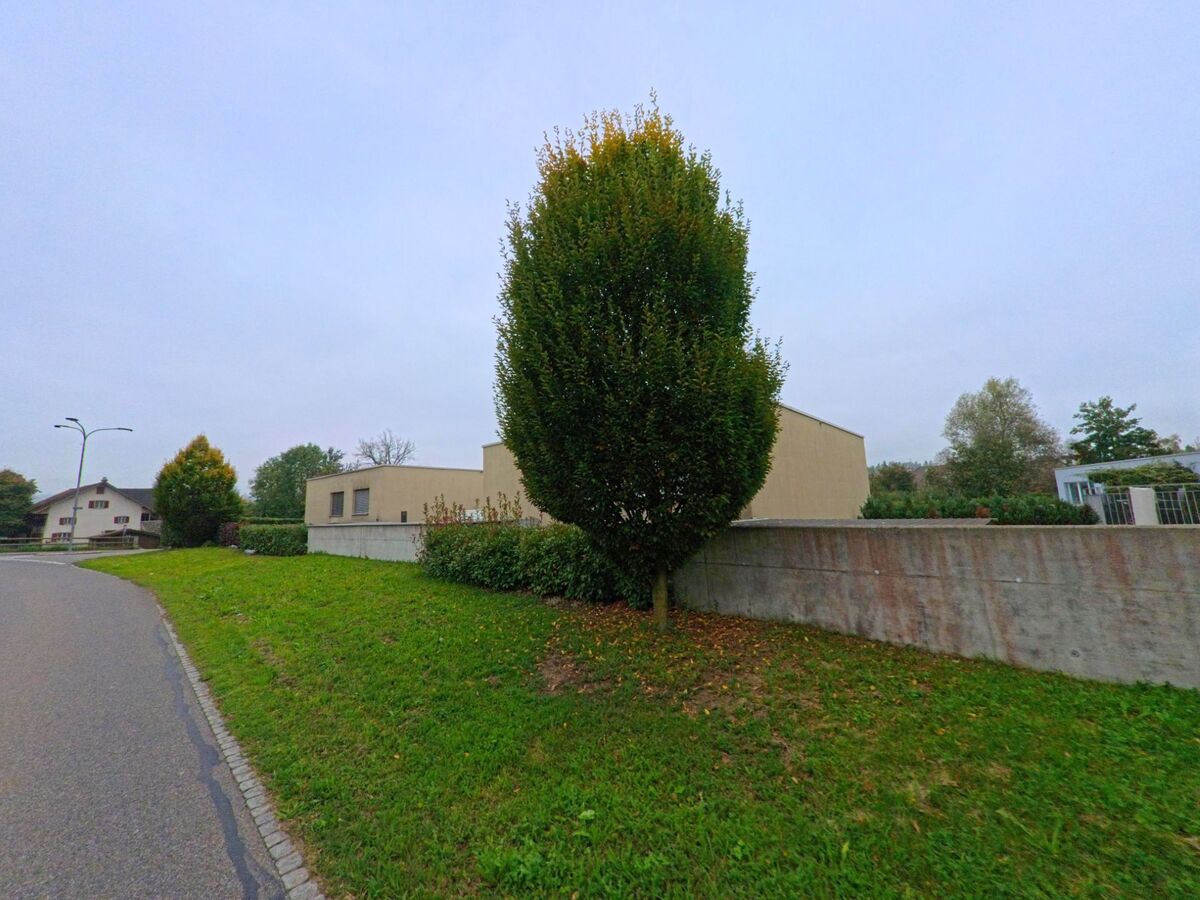 Eine Strasse in Stetten (5608, Kanton Aargau) fuehrt an einer niedrigen Betonmauer vorbei, hinter der ein einzelner Baum und moderne Gebaeude stehen.