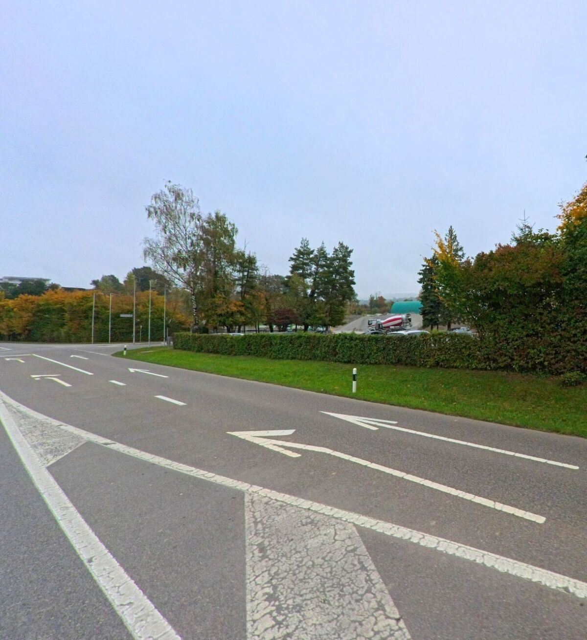 Eine mehrspurige Strasse in Stetten (5608, Kanton Aargau) fuehrt an einer dicht bewachsenen Hecke vorbei, hinter der sich ein Parkplatz mit Fahrzeugen befindet.
