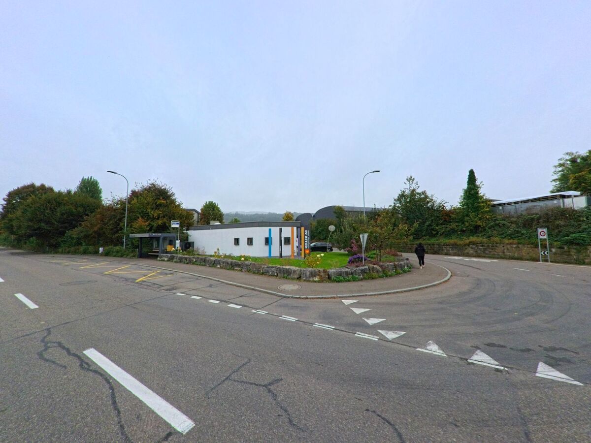 An einer kleinen Verkehrsinsel in Stetten (5608, Kanton Aargau) liegen eine Bushaltestelle und ein flaches Gebaeude, umgeben von Baeumen und Straeucher.
