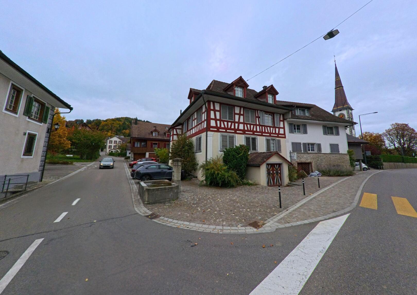 An einer ruhigen Strassenkreuzung in Stettfurt (9507, Kanton Thurgau) prägen traditionelle Fachwerkhäuser und der Kirchturm das dörfliche Ortsbild unter leicht bewölktem Himmel.