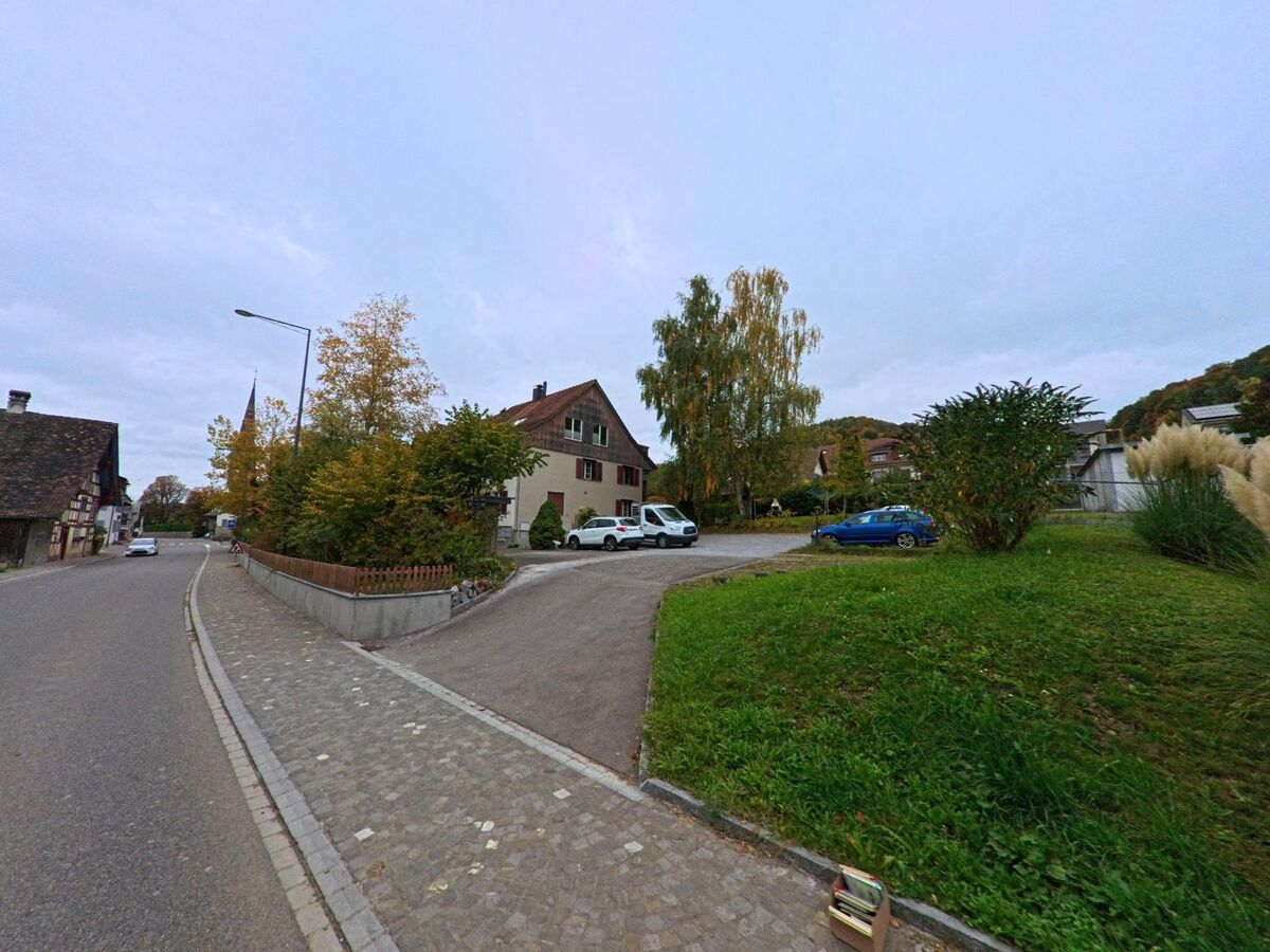 Neben einer leicht ansteigenden Zufahrt zeigt sich in Stettfurt (9507, Kanton Thurgau) ein Wohnhaus mit Parkplatz, umgeben von Gruenflaechen und Baeumen im herbstlichen Dorfumfeld.