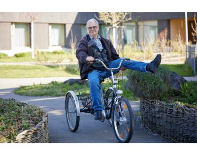 Ein Mann faehrt in Kloten (8302) im Kanton Zuerich mit einem Dreirad auf einem Weg vor einem Gebaeude der Stiftung Pigna.