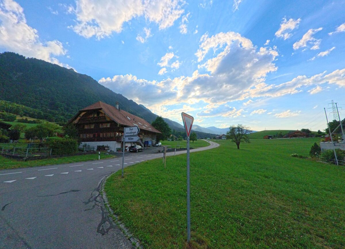 In Stocken-Hoefen (PLZ 3632, Kanton Bern) steht an einer Weggabelung mit Wegweisern ein grosses Bauernhaus vor bewaldetem Hang und weiter Felderlandschaft.
