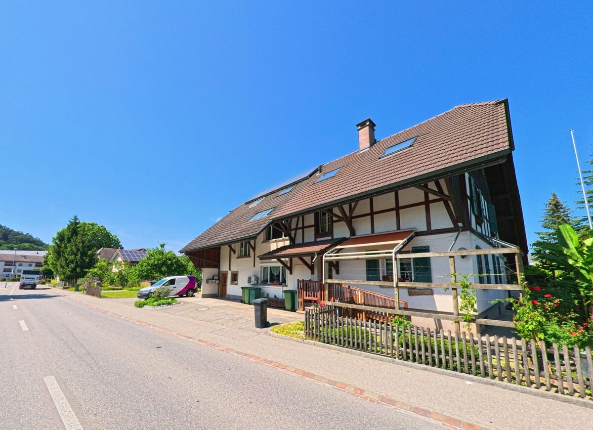 In Studen (2557, Kanton Bern) führt eine Strasse entlang eines traditionellen Fachwerkhauses, während moderne Häuser und Solarpaneele im Hintergrund sichtbar sind.