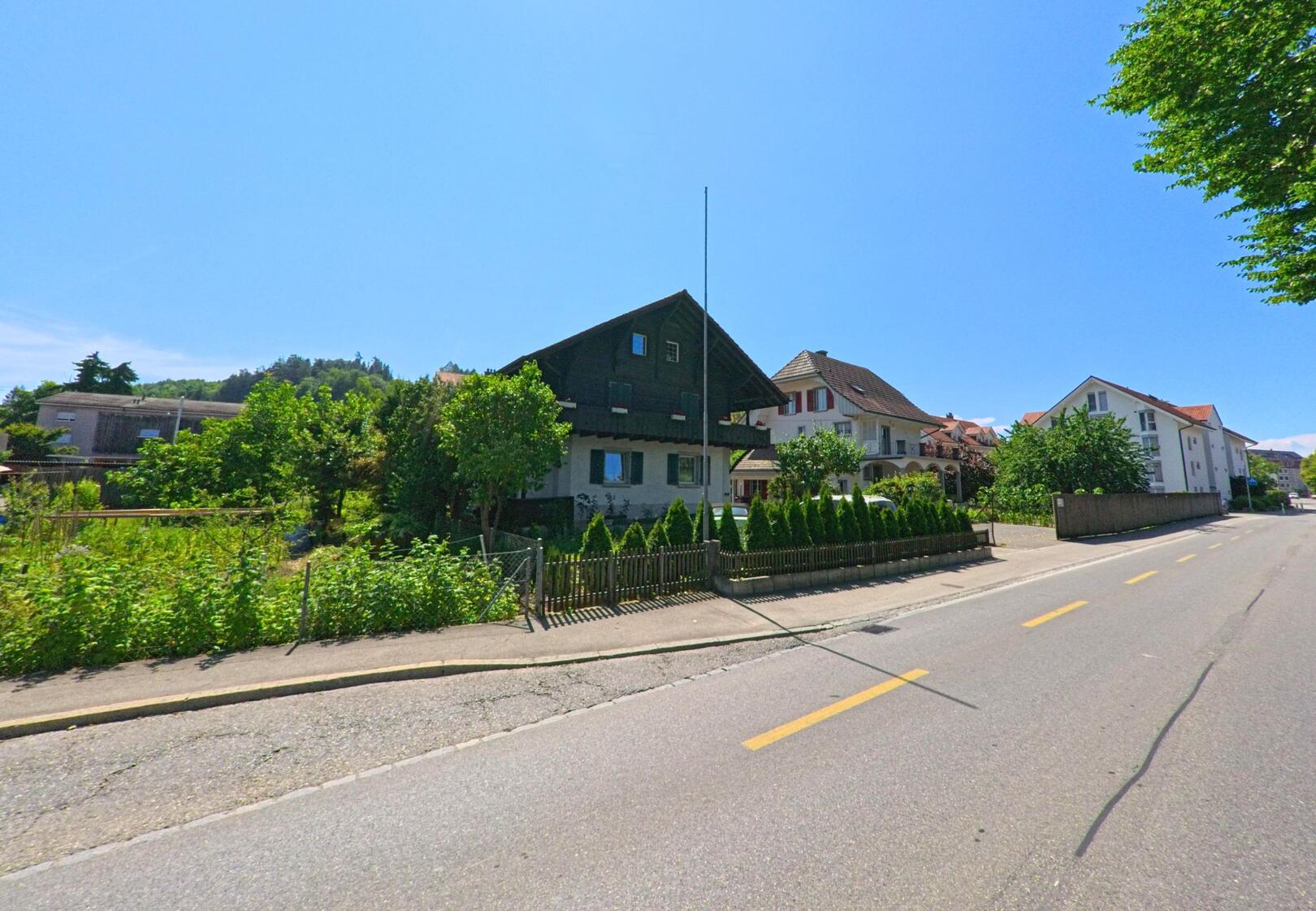 In Studen (2557, Kanton Bern) führt eine breite Strasse entlang gepflegter Häuser und grüner Gärten, mit einem sonnigen Himmel im Hintergrund.