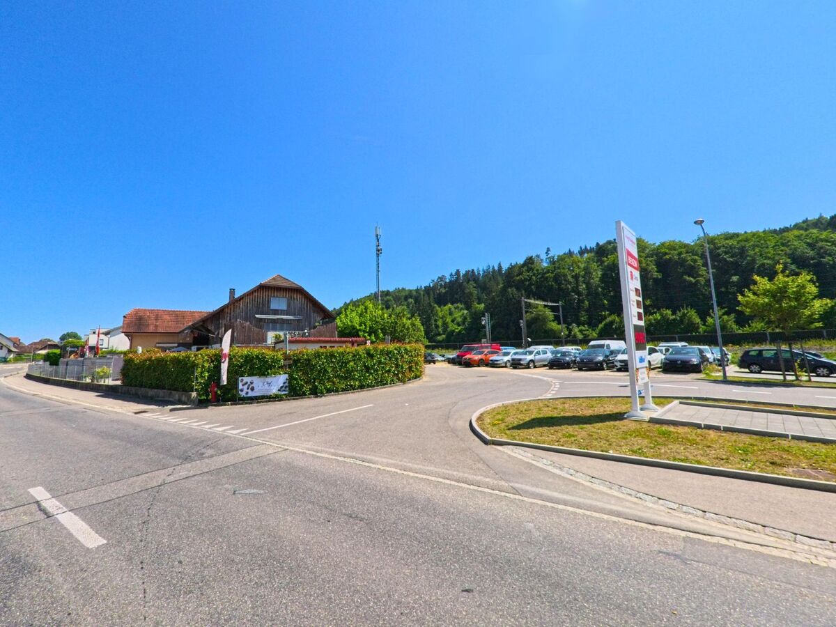 In Studen (2557, Kanton Bern) führt eine Strasse an einem traditionellen Gebäude und einem Parkplatz entlang, der von Bäumen und grünen Hügeln im Hintergrund umrahmt wird.
