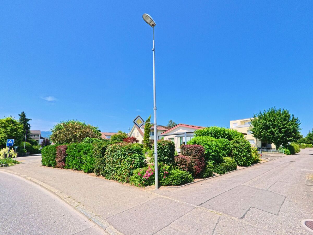 In Studen (2557, Kanton Bern) führt eine Strasse an gepflegten Gärten und Hecken entlang, mit einem Wegweiser und einer Straßenlaterne im Vordergrund.