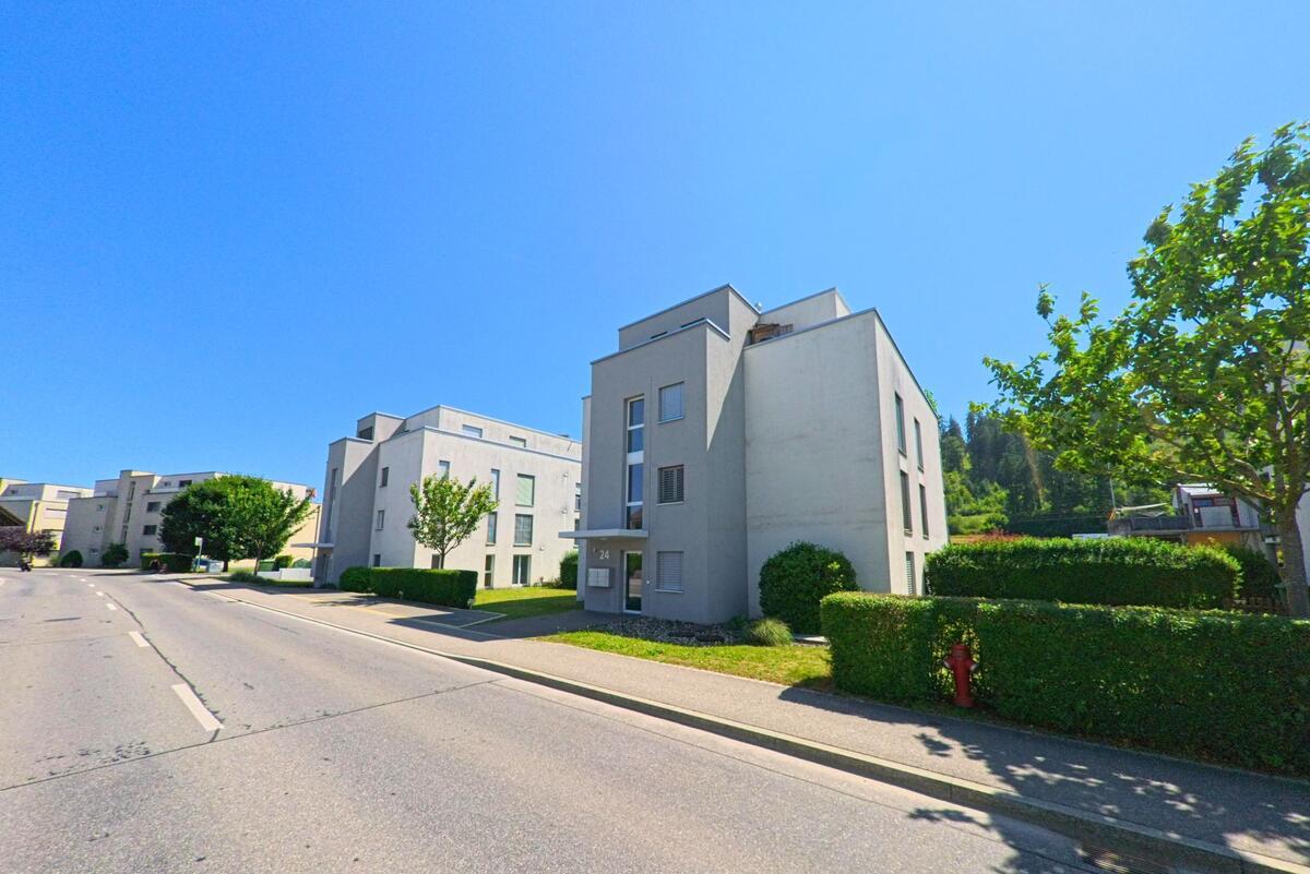 In Studen (2557, Kanton Bern) verläuft eine Strasse an modernen, funktionalen Wohngebäuden entlang, die von grünen Hecken und Bäumen umgeben sind.