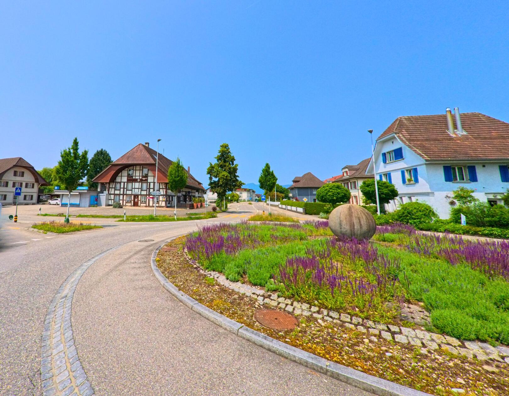 In Subingen (4553, Kanton Solothurn) befindet sich ein Kreisverkehr mit einer grossen Steinskulptur und begrünten Flächen, umgeben von traditionellen und modernen Häusern.