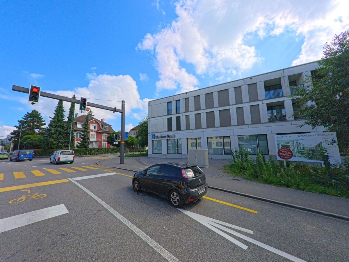 An einer Ampelkreuzung in Suhr (5034, Kanton Aargau) stehen moderne Geschaeftsgebaeude neben aelteren Wohnhaeusern mit hohen Nadelbaeumen.
