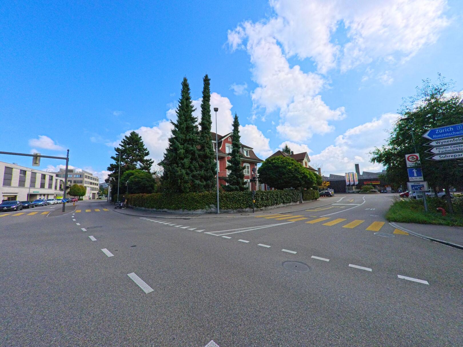 An einer vielbefahrenen Kreuzung in Suhr (5034, Kanton Aargau) stehen Wohnhaeuser mit hohen Nadelbaeumen, waehrend Wegweiser den Verkehr leiten.