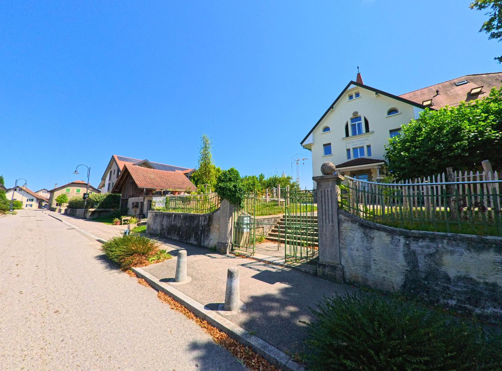 In Sullens (1036, Kanton Waadt) verlaeuft eine ruhige Strasse vorbei an traditionellen Wohnhaeusern und einem grossen Gebaeude mit Gartenanlage.
