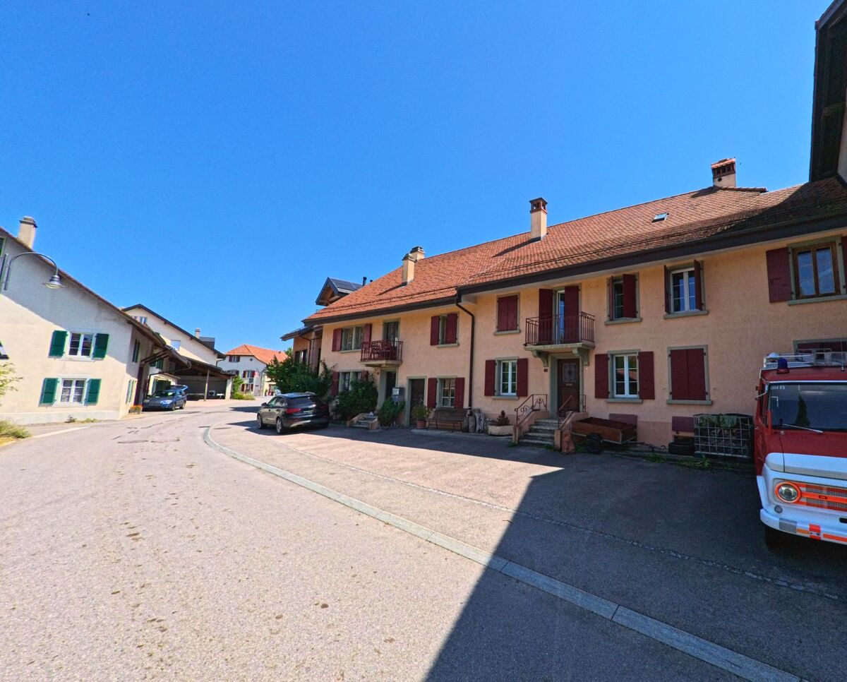 Eine leicht gekruemmte Dorfstrasse in Sullens (1036, Kanton Waadt) wird von traditionellen Wohnhaeusern mit roten und gruenen Fensterlaeden gesaeumt.