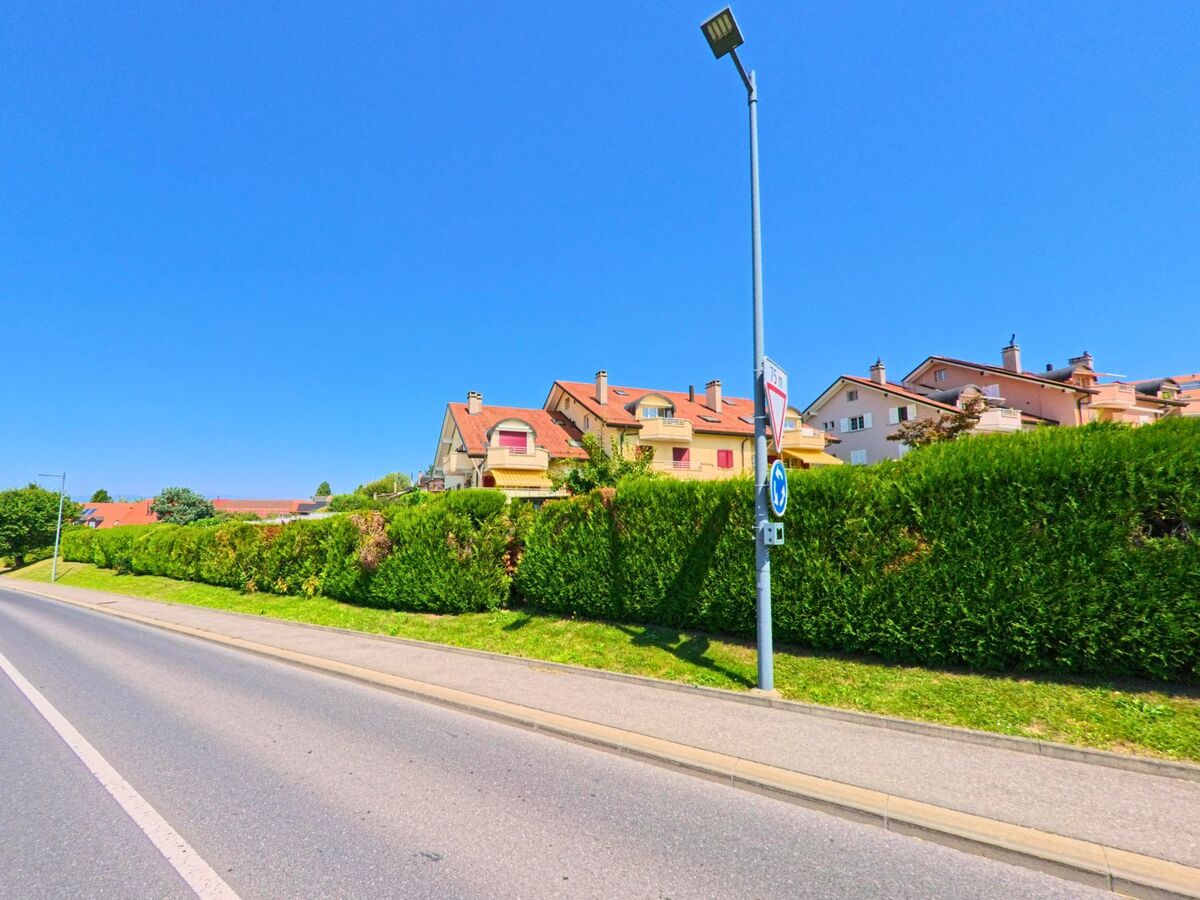 An einer Hauptstrasse in Sullens (1036, Kanton Waadt) stehen moderne Wohnhaeuser mit roten Daechern hinter einer hohen Hecke.