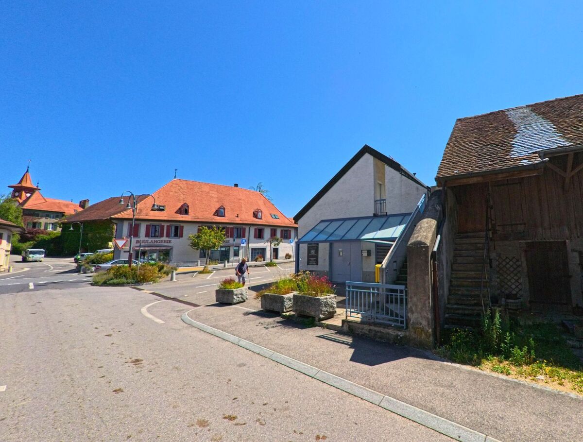 Im Zentrum von Sullens (1036, Kanton Waadt) stehen Wohnhaeuser mit roten Daechern, eine Baeckerei und ein kleiner Platz mit Fussgaengerwegen.