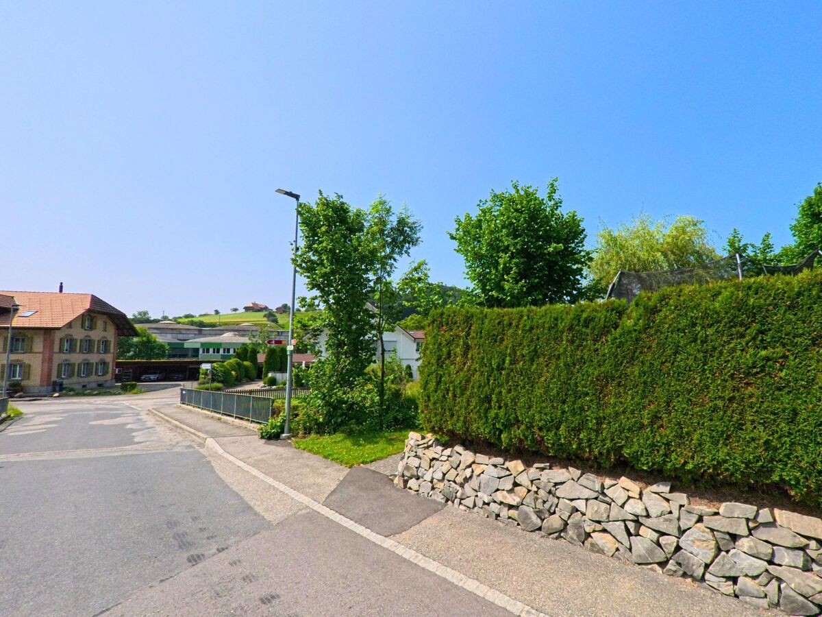 In Sumiswald (3454, Kanton Bern) führt eine Strasse an gepflegten Gärten und Bäumen entlang, mit einem Blick auf sanfte Hügel im Hintergrund.