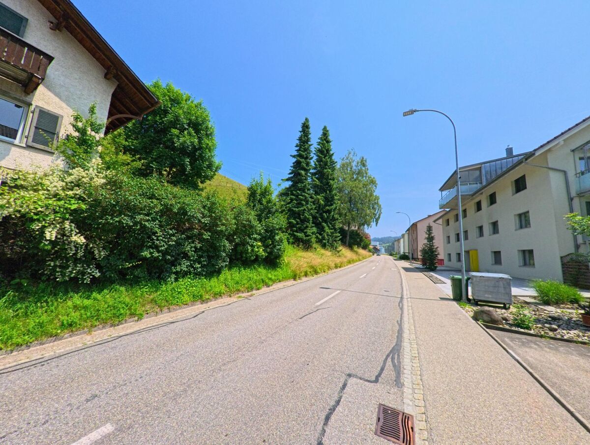 In Sumiswald (3455, Kanton Bern) führt eine ruhige Strasse zwischen gepflegten Häusern und Bäumen entlang, mit sanften Hügeln im Hintergrund.