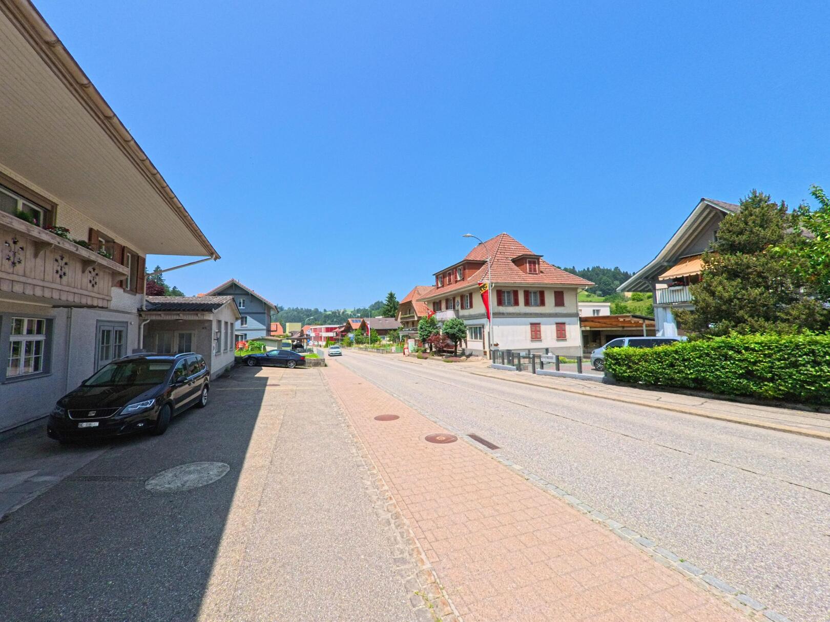 In Sumiswald (3457, Kanton Bern) führt eine ruhige Strasse zwischen historischen Gebäuden, mit parkenden Autos und grünen Sträuchern entlang der Wege.