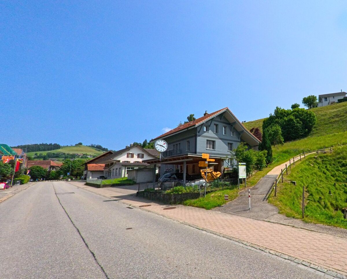 In Sumiswald (3457, Kanton Bern) führt eine Strasse entlang von Häusern, mit einem markanten Uhrenturm und einem Hügel im Hintergrund, der von einem Wanderweg flankiert wird.