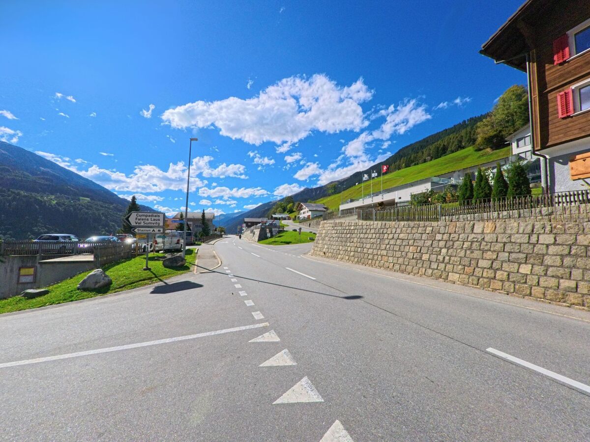 Am Ortseingang von Sumvitg (7175, Kanton Graubünden) führt eine breite Strasse mit Wegweisern durch das Tal, begleitet von Stützmauern, Wiesen und Hanglagen.