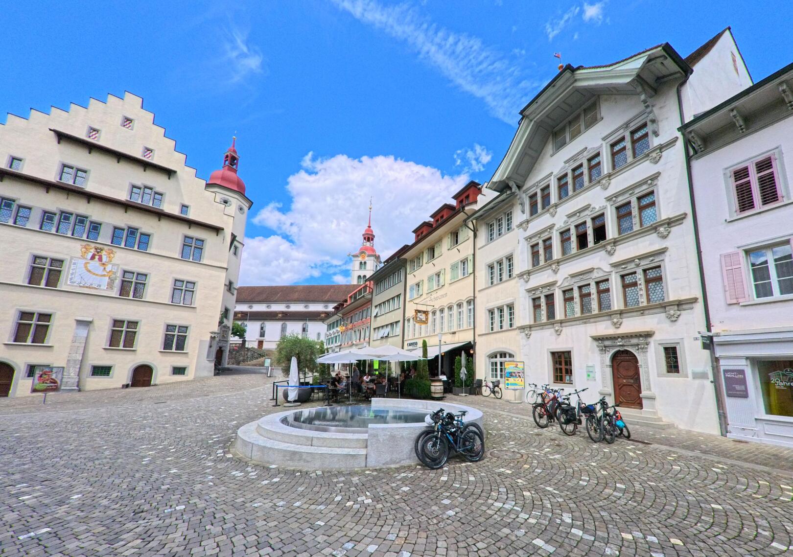 Auf einem gepflasterten Platz mit historischen Gebaeuden und Brunnen sitzen Menschen in Sursee (6210, Kanton Luzern) bei sonnigem Wetter.
