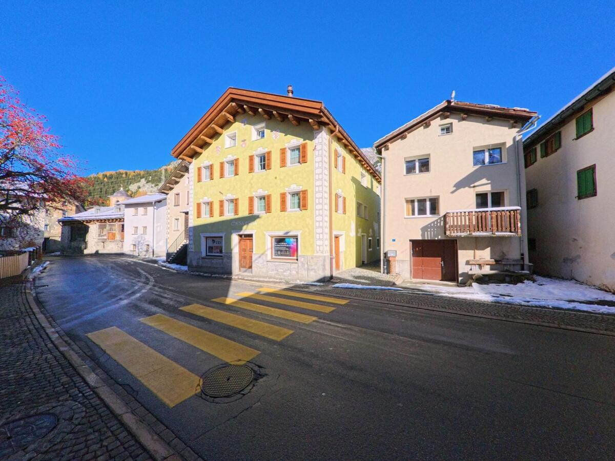 An einer übersichtlichen Kreuzung in Surses (7457, Kanton Graubünden) stehen farbig gefasste Wohnhäuser entlang einer Dorfstrasse mit Fussgängerstreifen und vereinzelten Schneeresten.