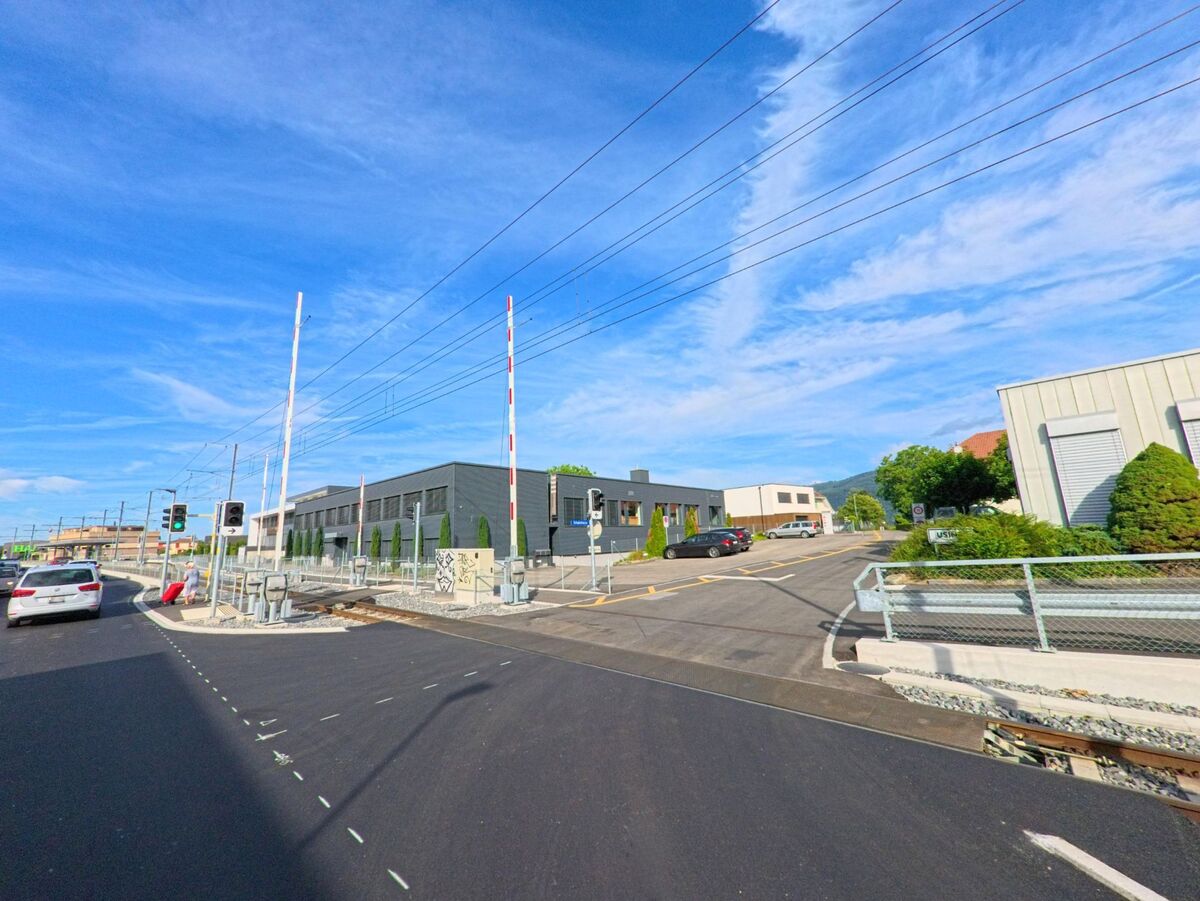 Ein Bahnübergang mit modernen Gebäuden und klarer Verkehrsführung befindet sich in Sutz-Lattrigen (2572, Kanton Bern) unter blauem Sommerhimmel.
