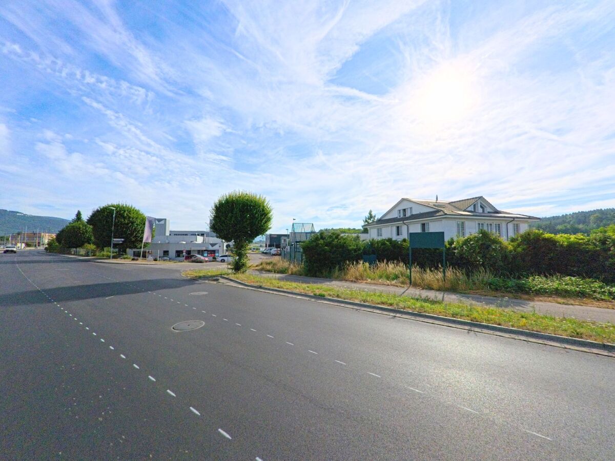 Am Ortseingang von Sutz-Lattrigen (2572, Kanton Bern) verläuft eine breite Strasse mit modernen Gebäuden und gepflegten Bäumen unter hellem Himmel.