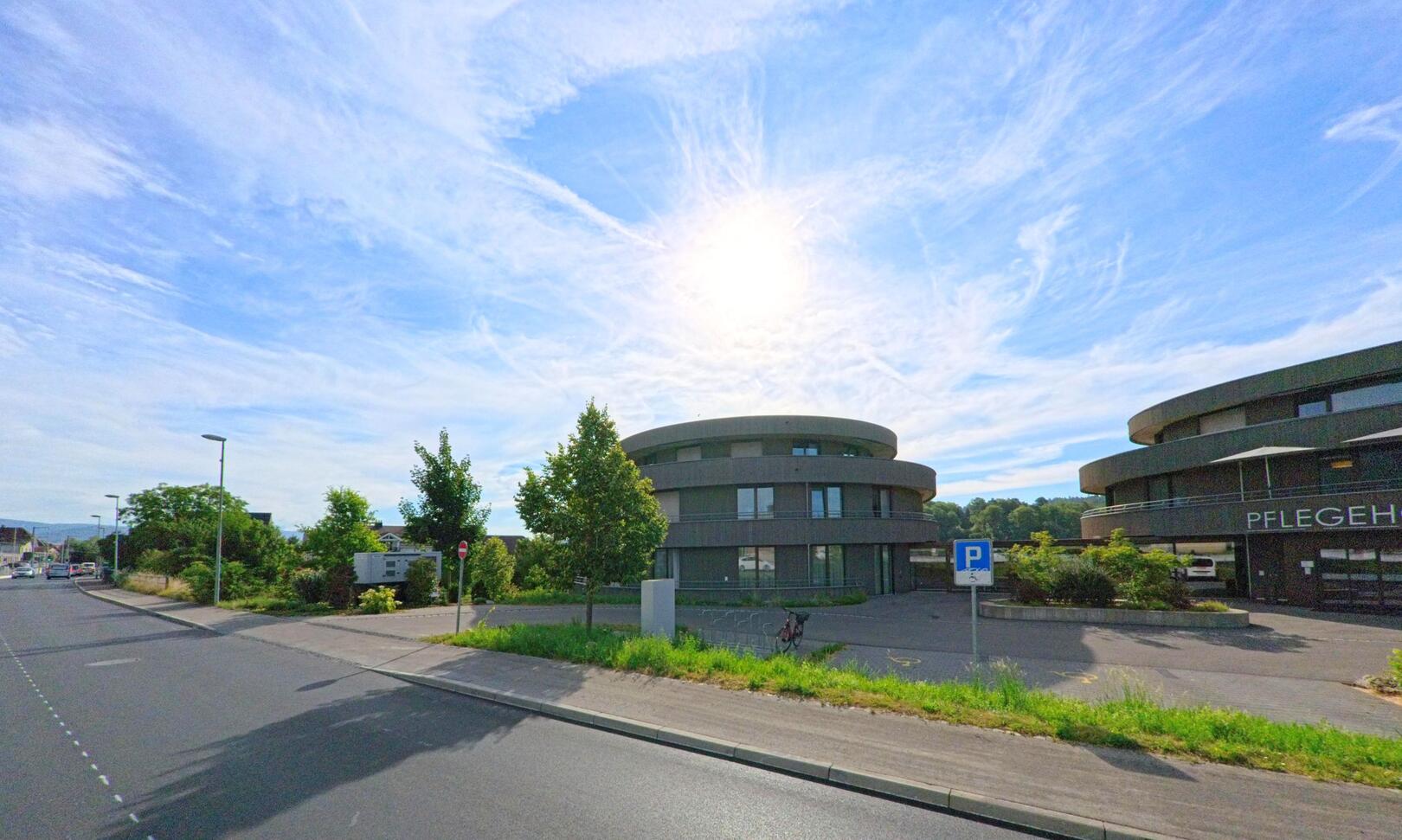 Runde moderne Gebäude des Pflegeheims stehen in Sutz-Lattrigen (2572, Kanton Bern) an einer breiten Strasse unter strahlendem Himmel.